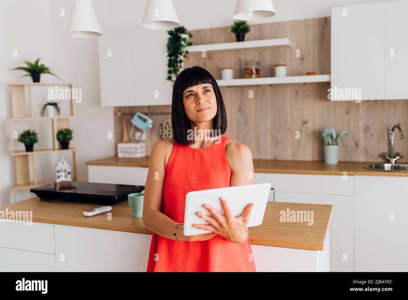 Thoughtful woman with tablet PC standing in kitchen at home Stock Photo