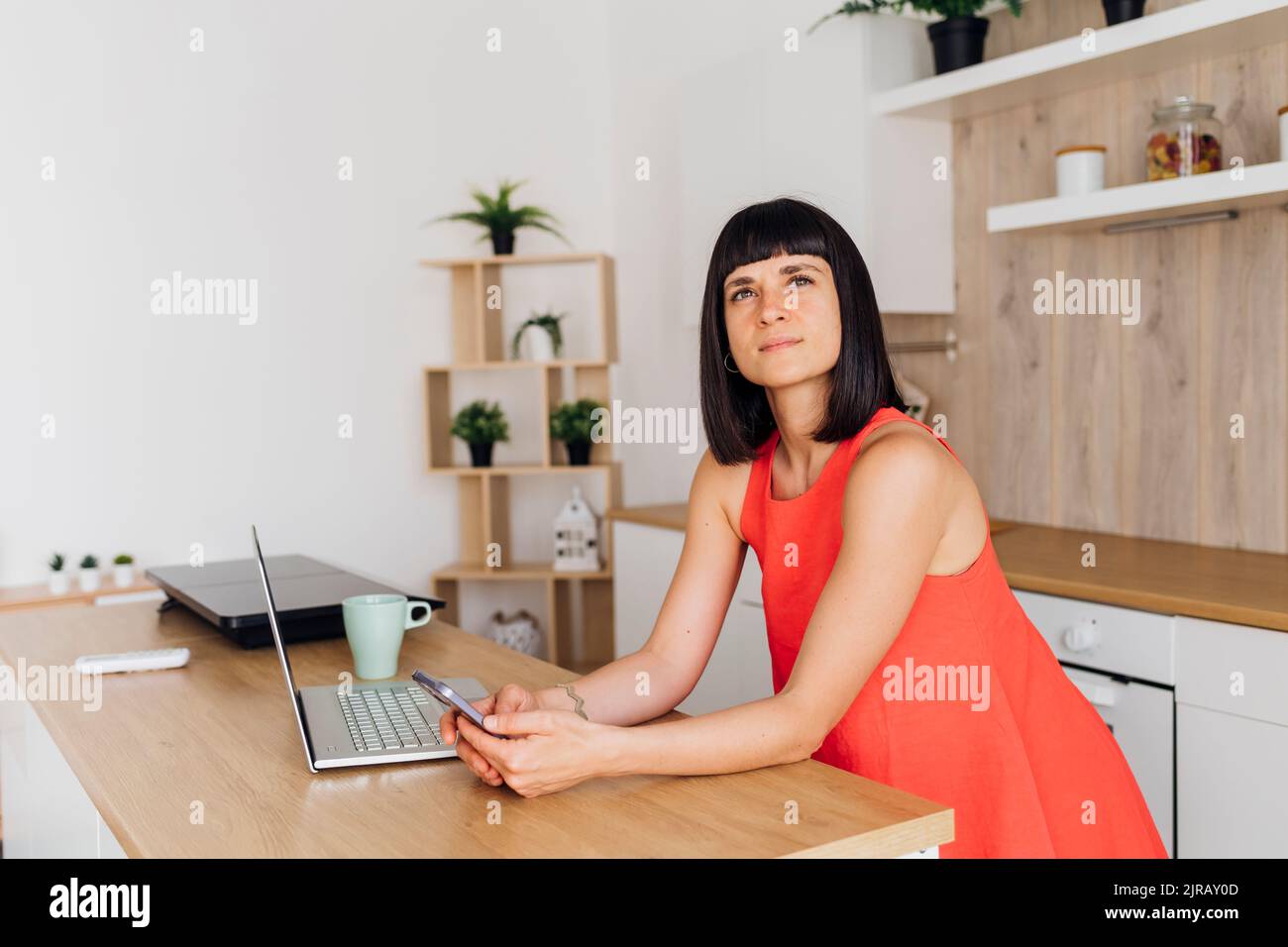 Contemplative woman with mobile phone and laptop standing at kitchen island Stock Photo