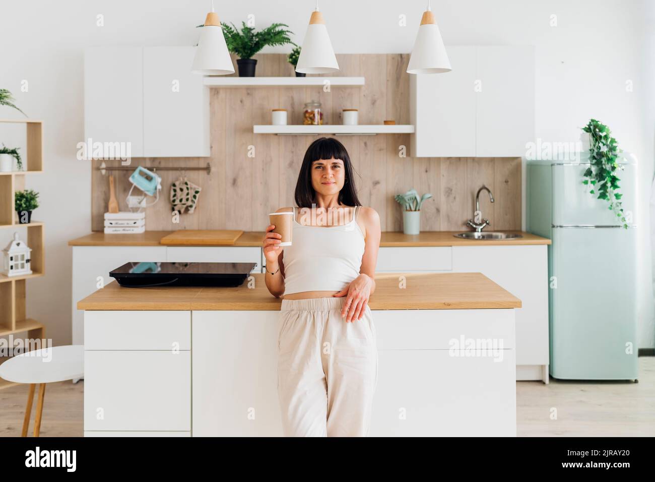 Woman holding disposable cup leaning on kitchen island at home Stock Photo