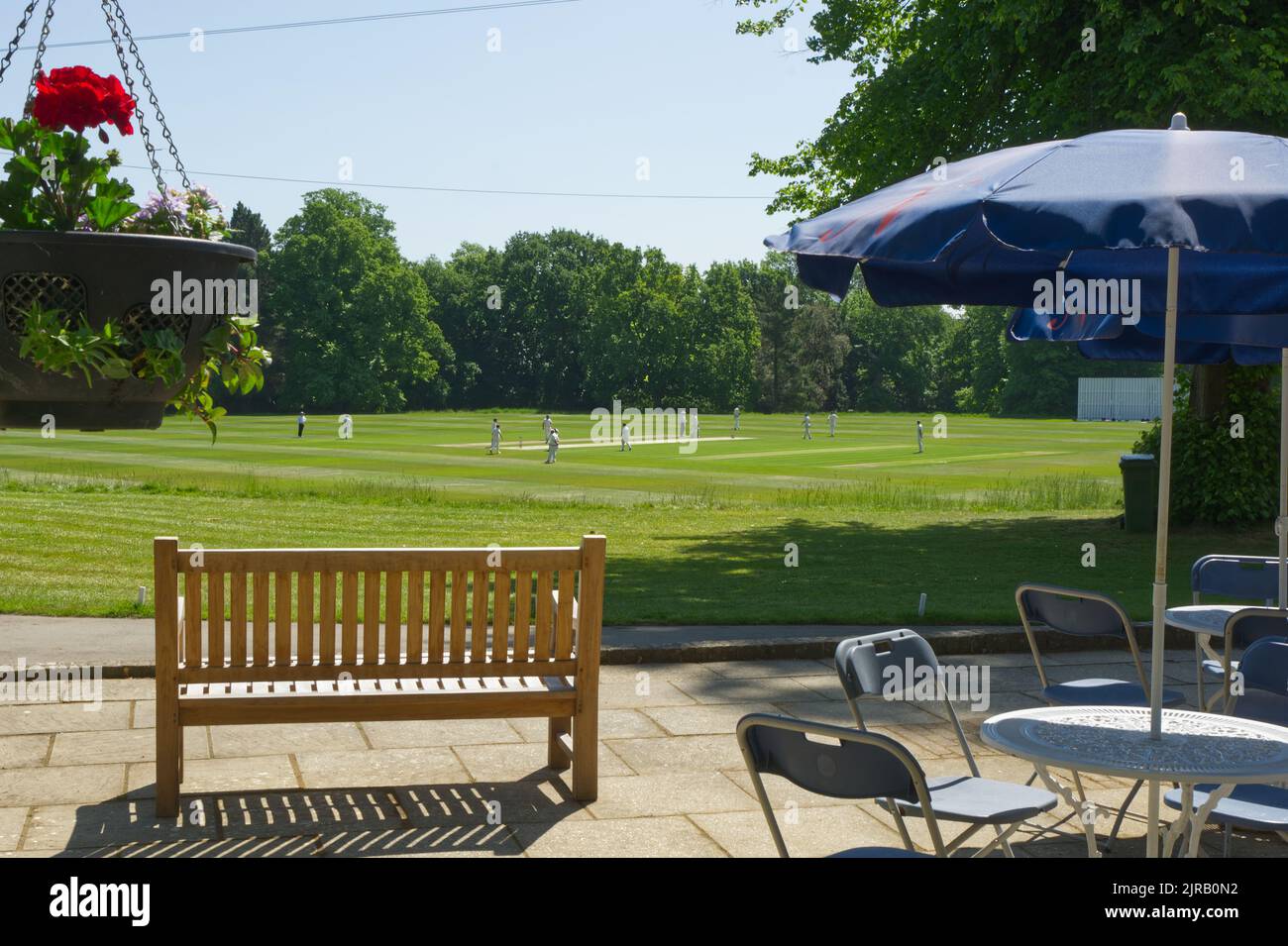 Cricket ground in rural setting with players on field and patio with bench and table and chairs. Unrecognisable people. Stock Photo