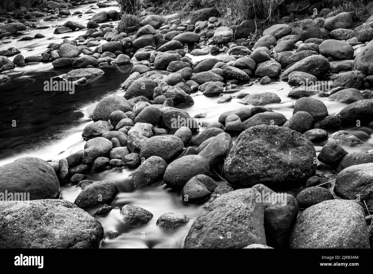 Long exposure of a fast flowing river between rounded basalt boulders in the Drakensberg Mountains of South Africa in black and white. Stock Photo