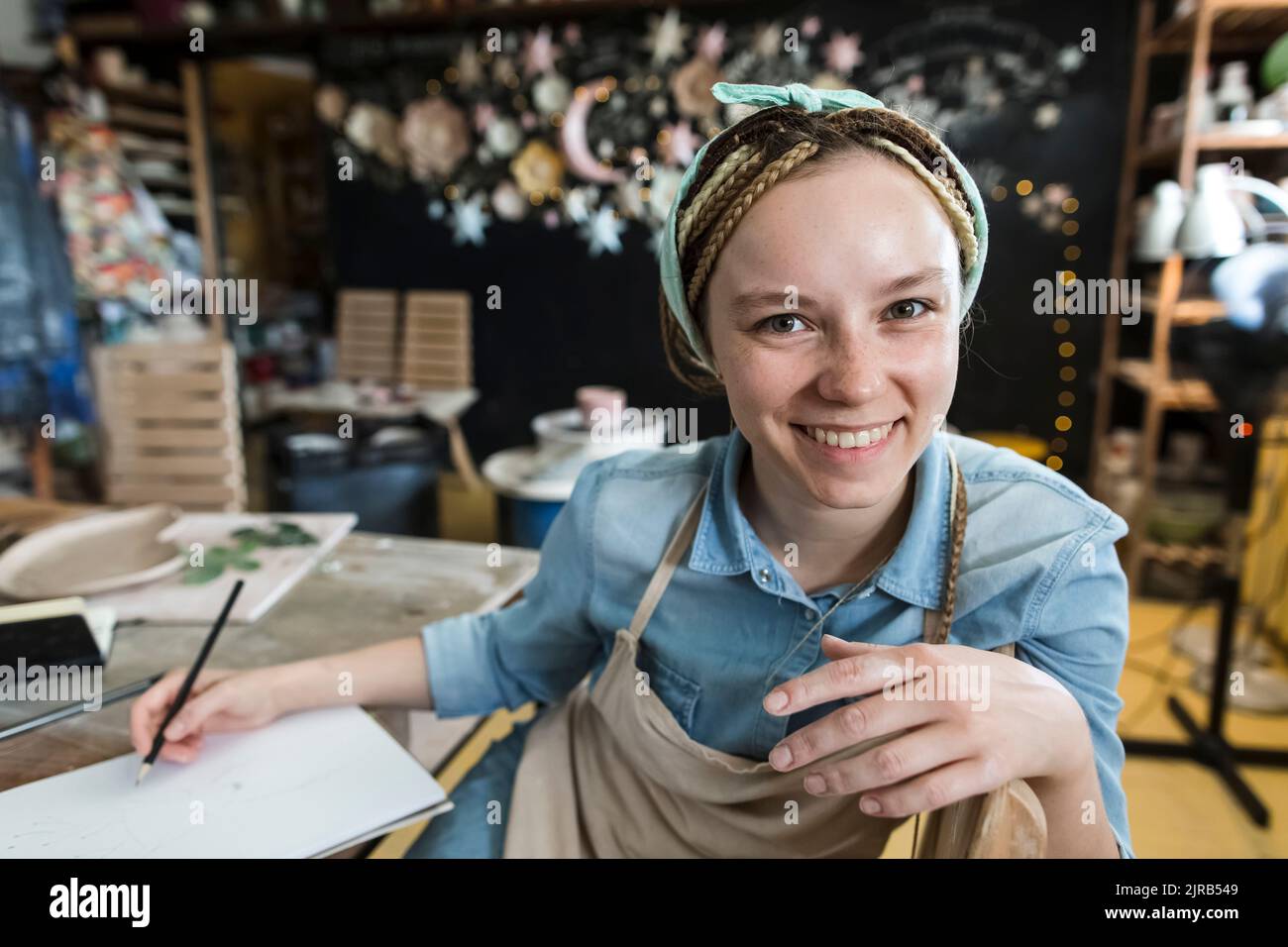 Happy craftsperson holding pencil sitting at workbench in art studio