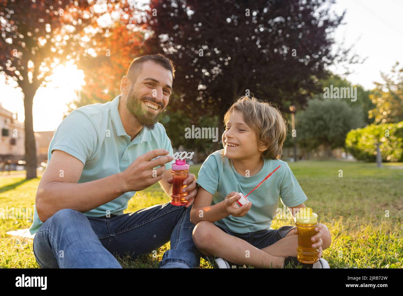 Smiling father and son with bubble wands sitting at park Stock Photo