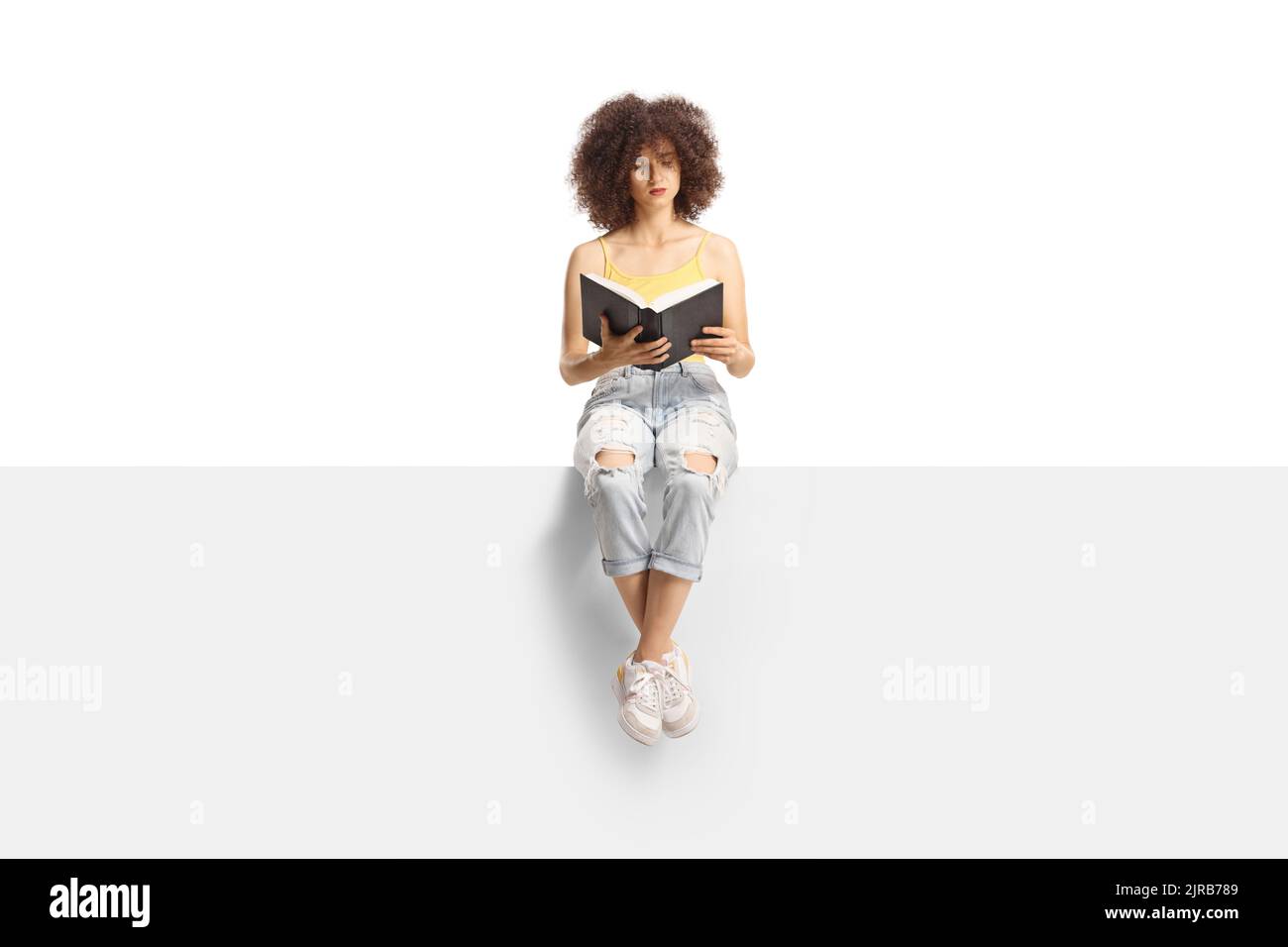 Young woman with curly hair sitting on a panel and reading a book isolated on white background Stock Photo