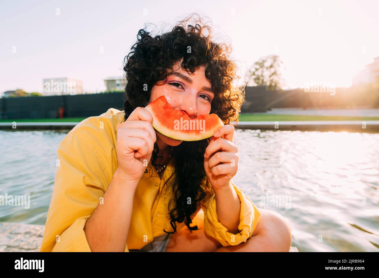 Young woman eating watermelon by pond at sunset Stock Photo