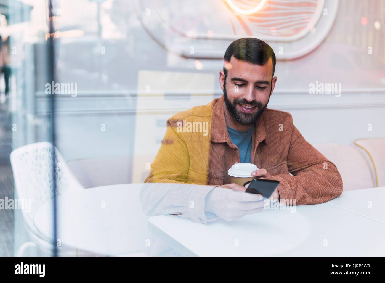 Smiling young man using smart phone seen through glass at cafe Stock Photo