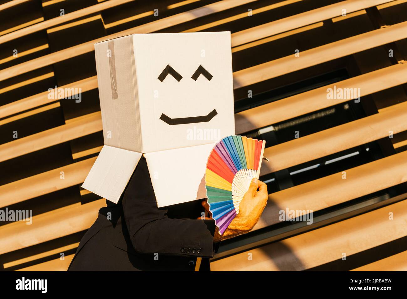 Businessman wearing box with smiley face holding rainbow color hand fan on sunny day Stock Photo