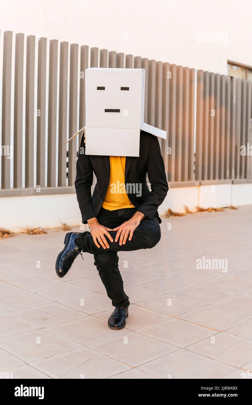Man wearing box with face balancing with legs crossed at knee on footpath Stock Photo