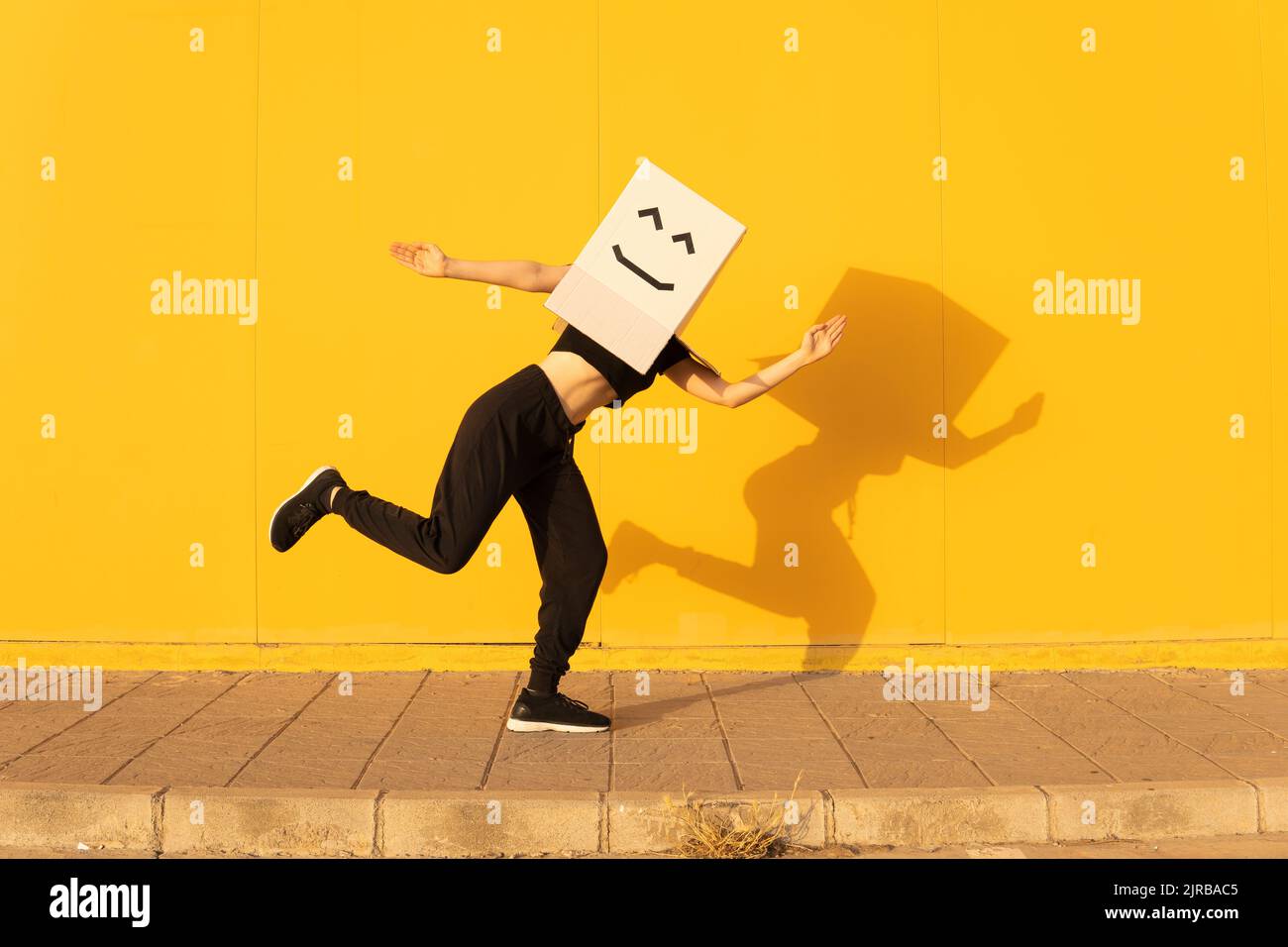 Playful woman wearing box with smiley face doing robot dance in front of yellow wall Stock Photo