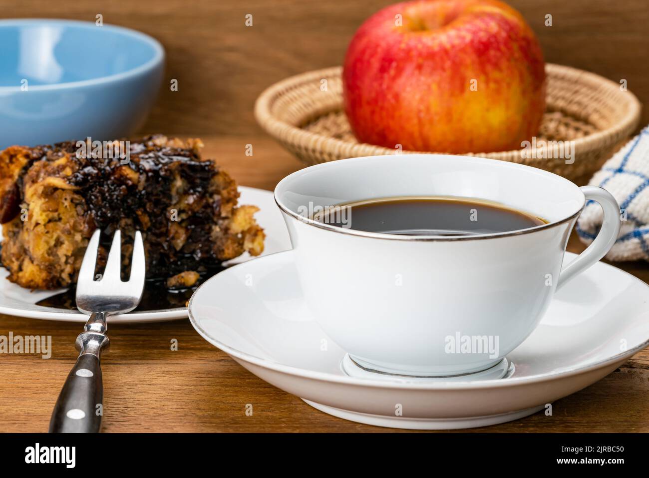 Black coffee in white ceramic cup on saucer with a portion of fruitcake poured with chocolate syrup in white ceramic dish, metal fork and a ripe red a Stock Photo