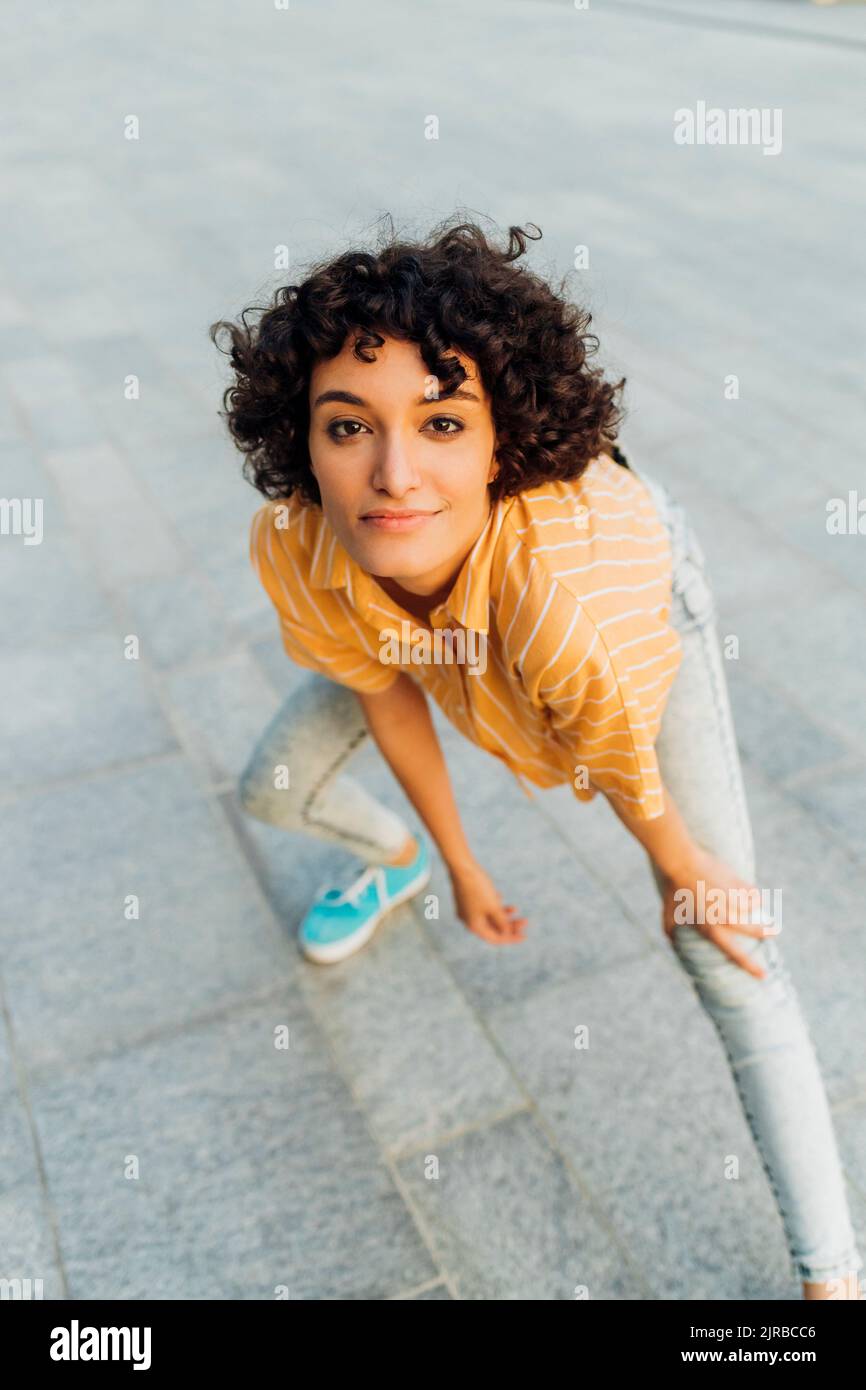 Smiling young woman with hand on knee standing on footpath Stock Photo