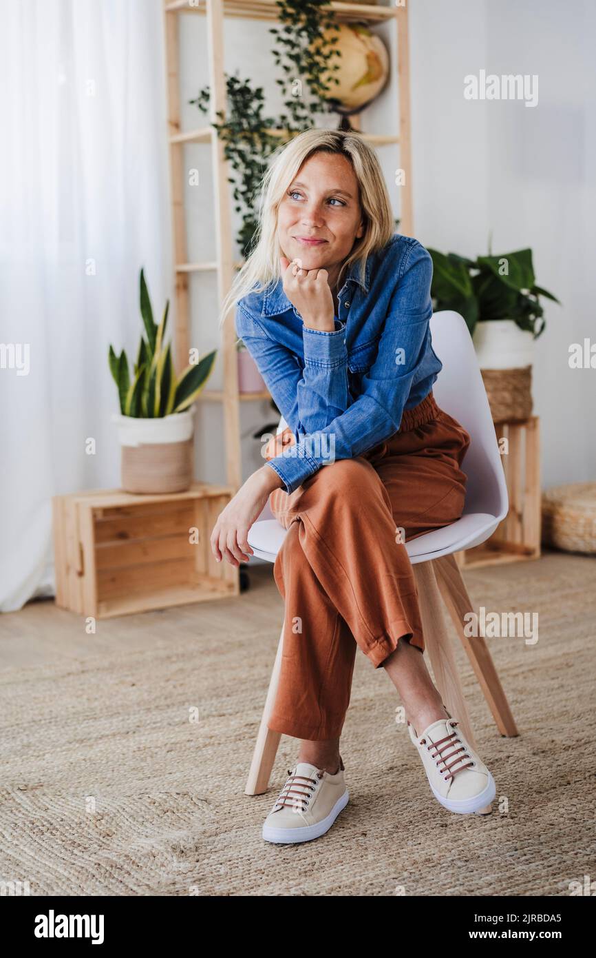 Contemplative blond woman with hand on chin sitting at home Stock Photo ...