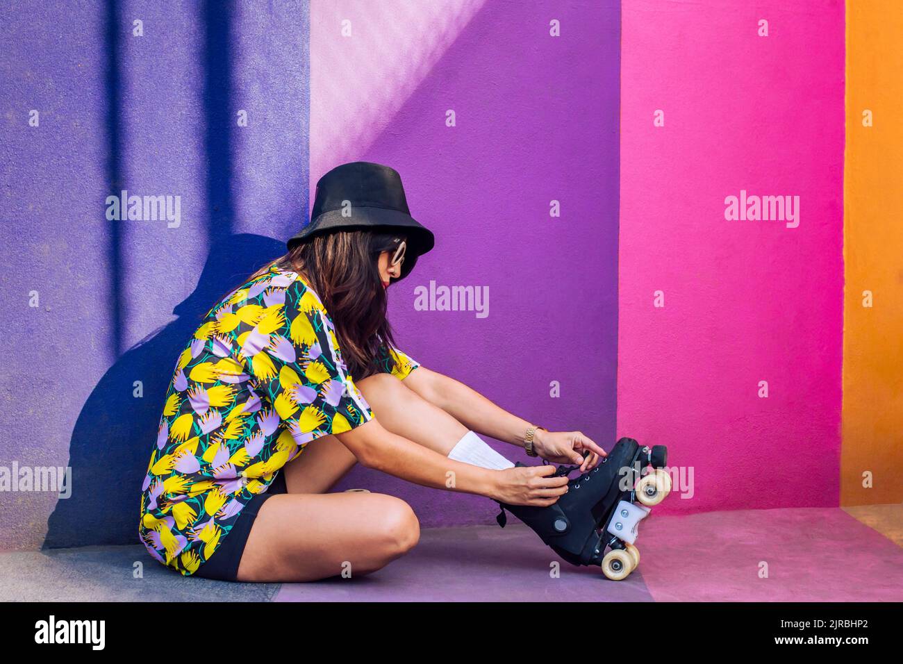 Woman wearing hat tying shoelace on roller skate by multi colored wall Stock Photo