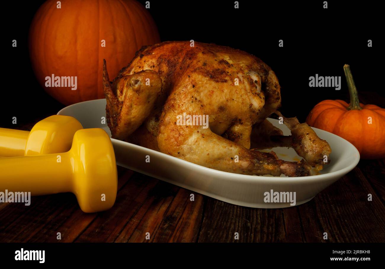 Roasted homemade chicken for Thanksgiving dinner, with pumpkins and two gym dumbbells. Autumn composition, healthy fitness lifestyle, diet concept. Stock Photo
