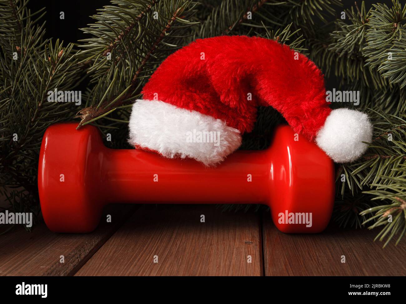 Red dumbbell with Santa Claus hat. Exercise equipment as Christmas gift idea. Gym fitness holiday season concept composition with tree branches. Stock Photo
