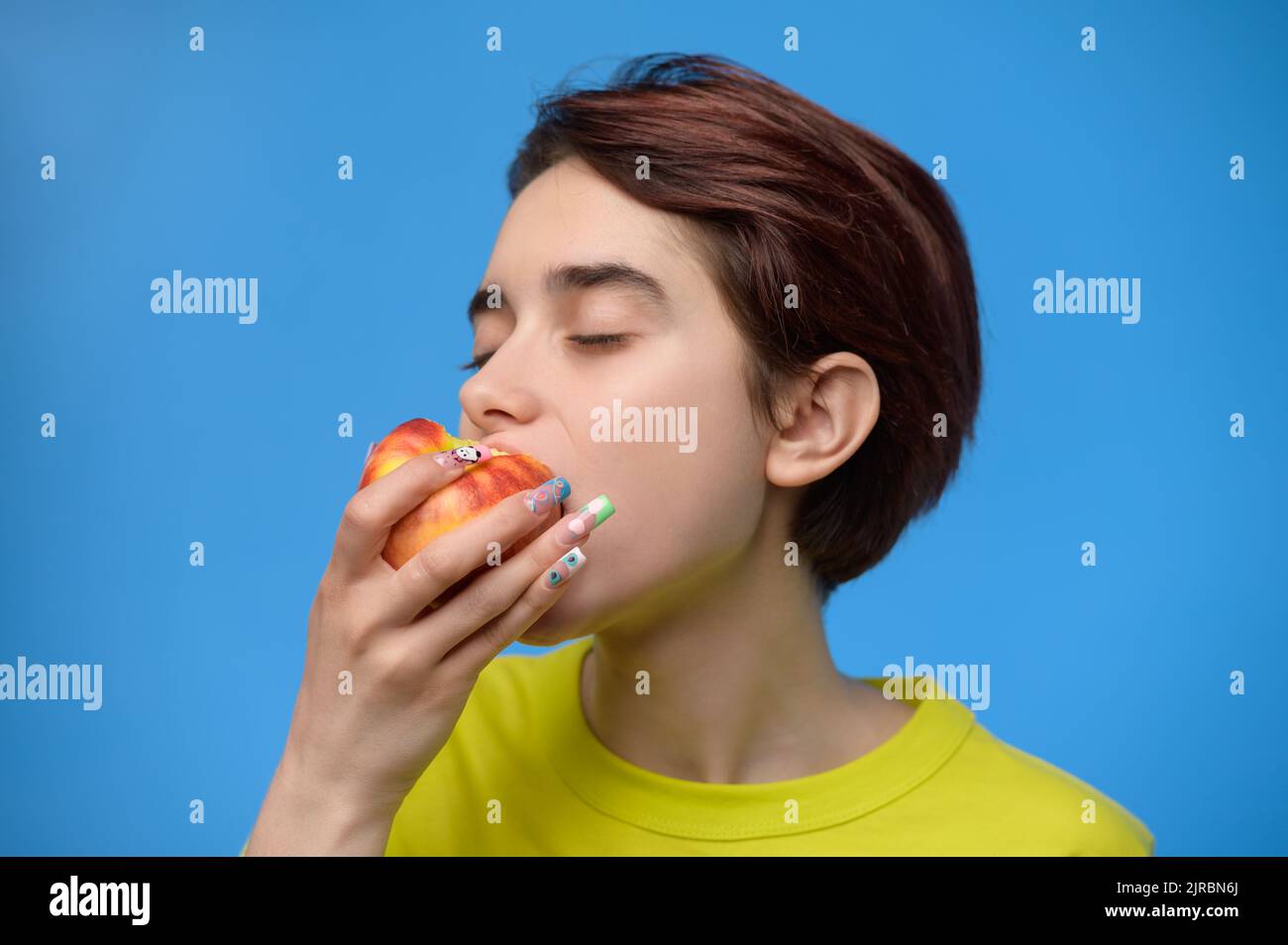 Close up portrait of pretty teenage girl biting an apple on blue background Stock Photo
