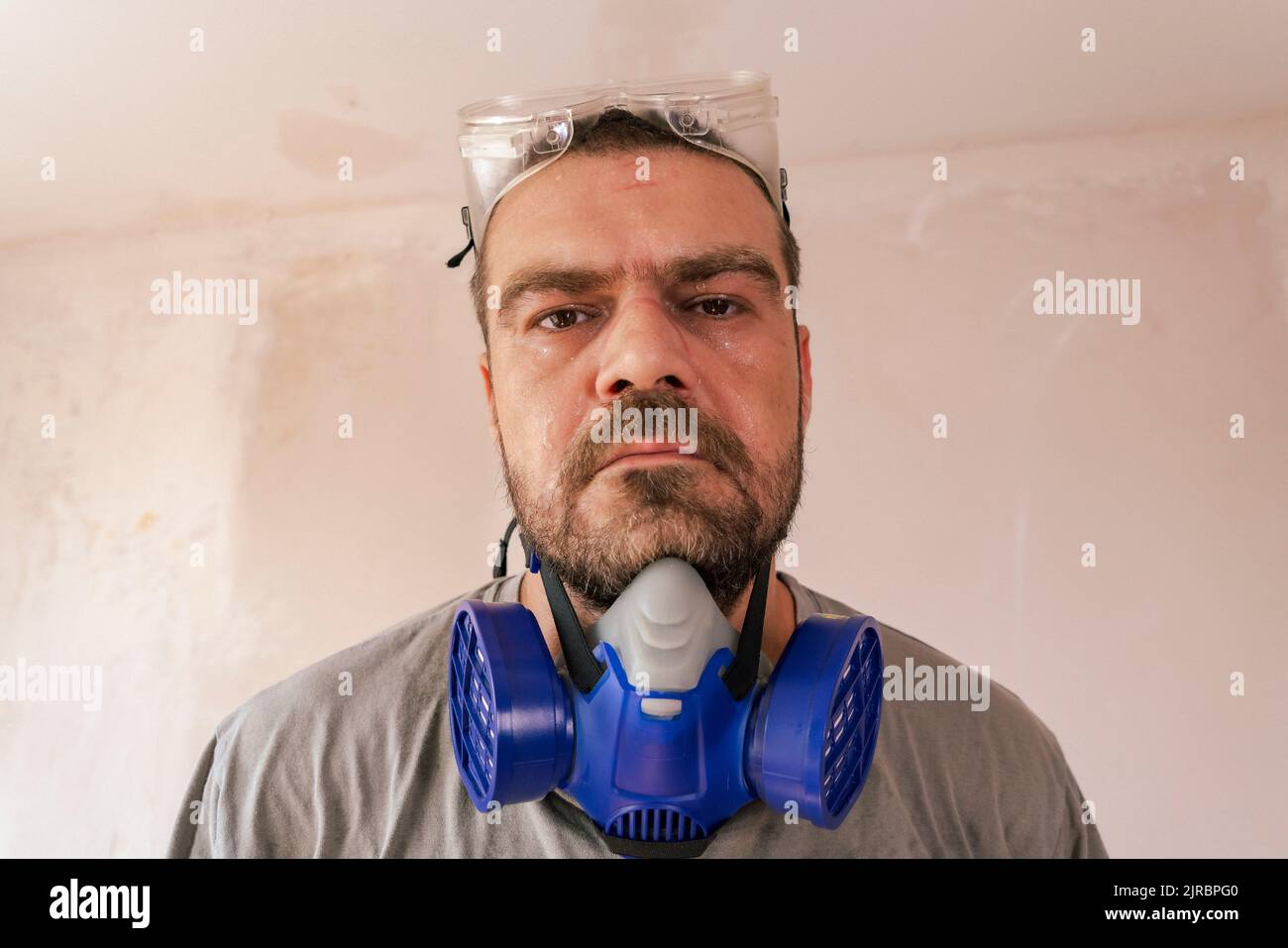 Selective focus shot of sweaty caucasian man with safety glasses and dust mask, looking directly into the lens seriously. Stock Photo