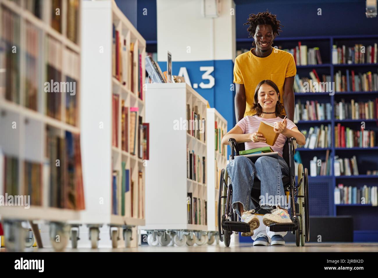 Vibrant full length shot of black young man assisting female student with disability in library and pushing wheelchair, copy space Stock Photo