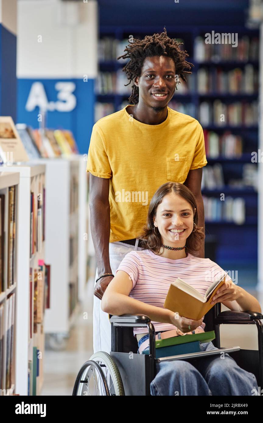 Vertical portrait of smiling young woman with disability looking at camera in college library with friend assisting Stock Photo