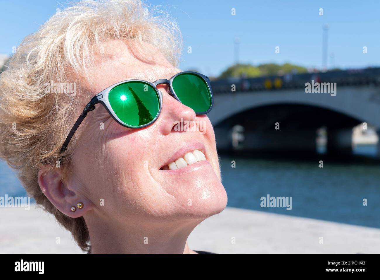 A female tourist visiting Paris, France looks up at the Eiffel Tower. The famous landmark is reflected in the woman’s Oakley prescription sunglasses Stock Photo