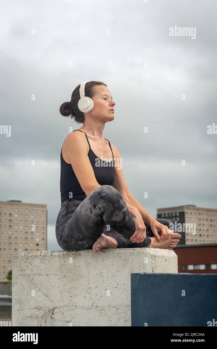 Woman sitting crossed legged wearing headphones listening to music outside, urban setting. Stock Photo