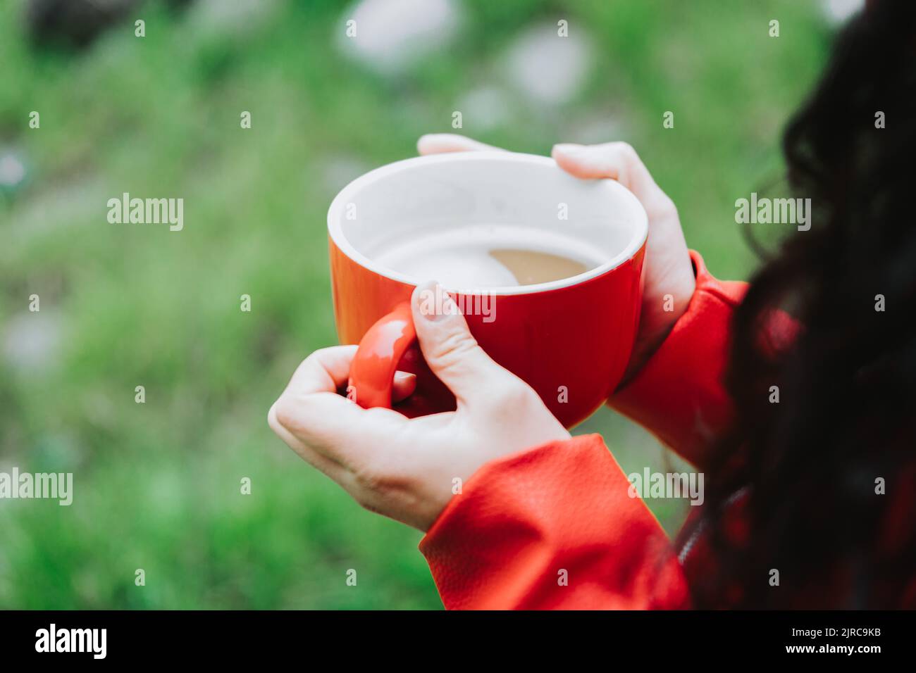 Unrecognizable person, wearing a red leather jacket, and holding a red mug with hot coffee in nature. Selective focus Stock Photo