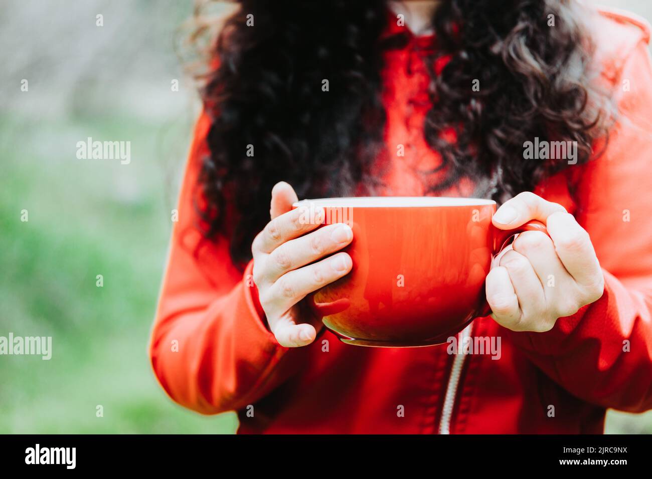 Curly brunette woman, wearing a red leather jacket, and holding a red mug with hot coffee in nature. Selective focus Stock Photo