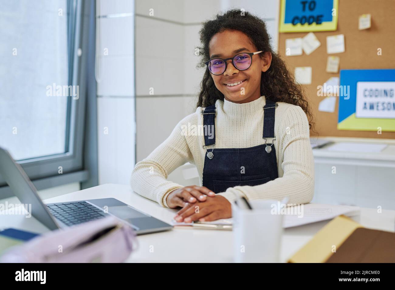 Portrait of smiling black schoolgirl sitting at desk with laptop in ...
