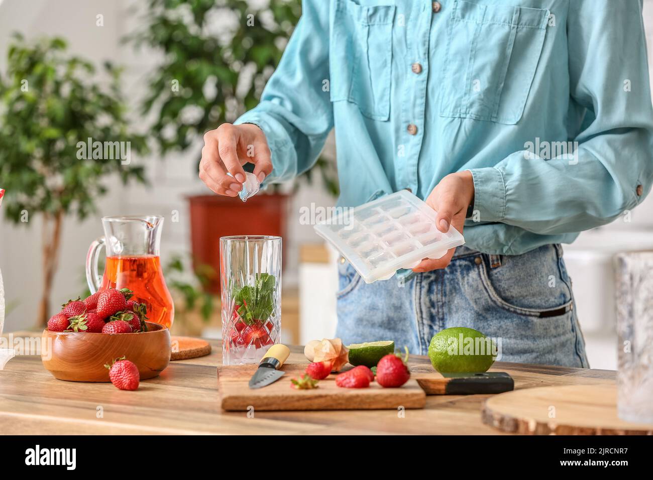 Woman putting ice cubes into glass for strawberry lemonade in kitchen