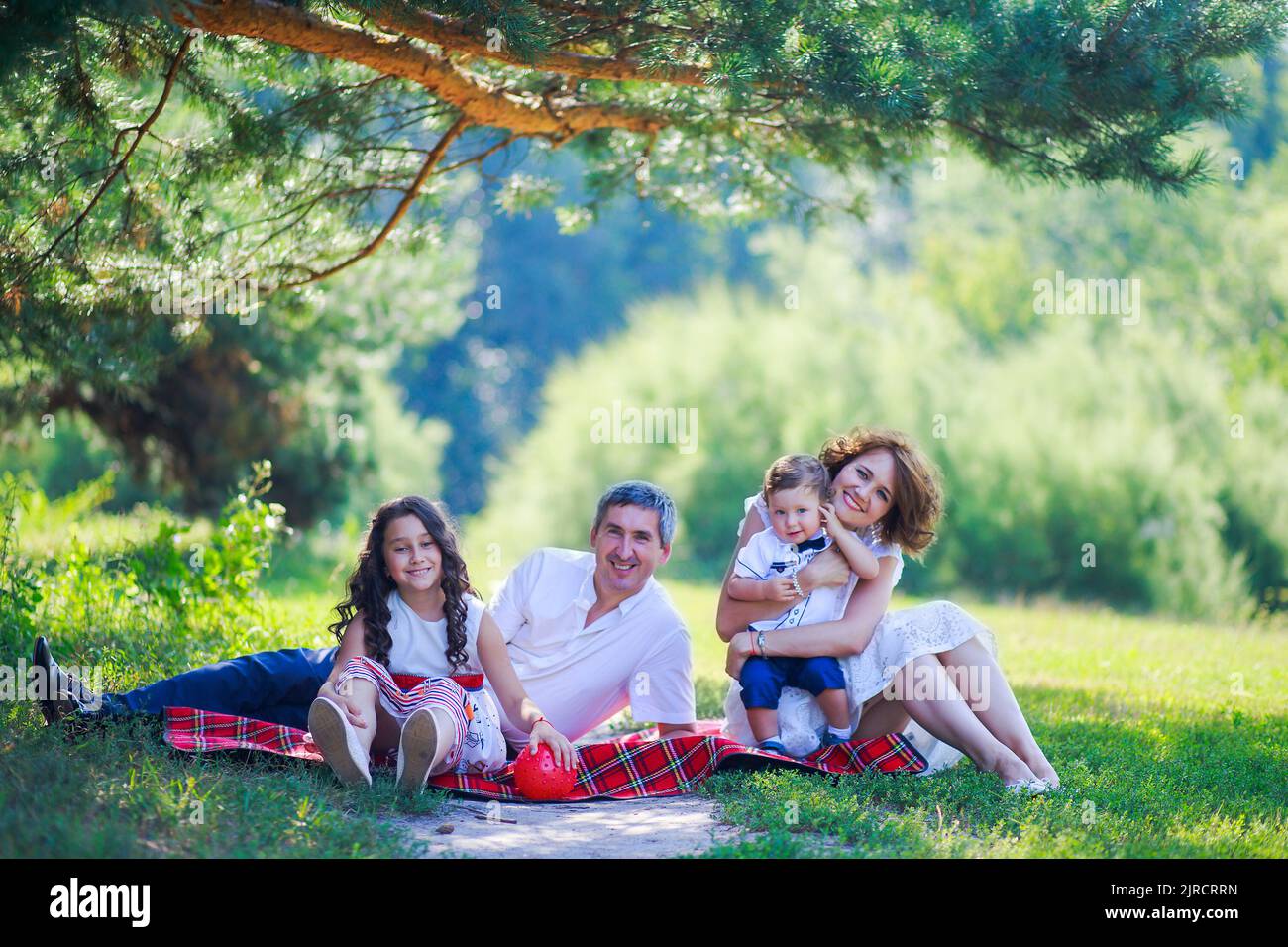 Young Family With Two Kids Sitting On The Grass Under A Pine Tree young-family-with-two-kids-sitting-on-the-grass-under-a-pine-tree