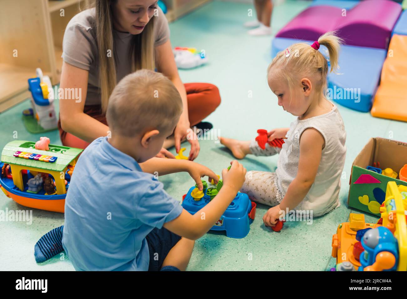Learning through sensory play at the nursery school. Caucasian toddlers and their teacher playing with a colorful plastic shape sorter set, playhouses, building blocks, cars and boats. Imagination, creativity, fine motor and gross motor skills development. High quality photo Stock Photo