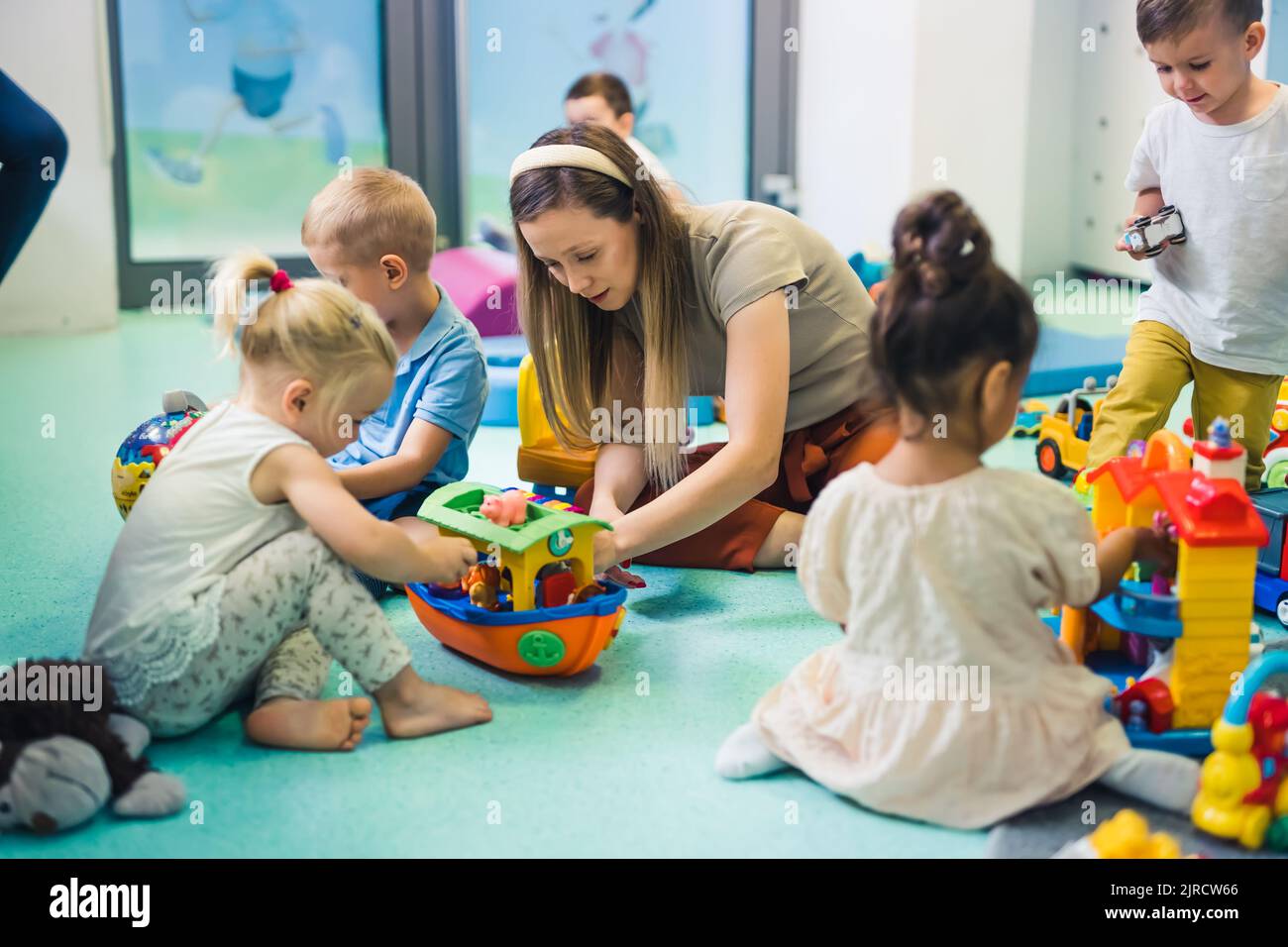Toddlers and their tutor playing with different colorful plastic toys, cars and boats while sitting on the floor in a playroom at the nursery school. Imagination, mathematical ability, fine motor and gross motor skills development. . High quality photo Stock Photo