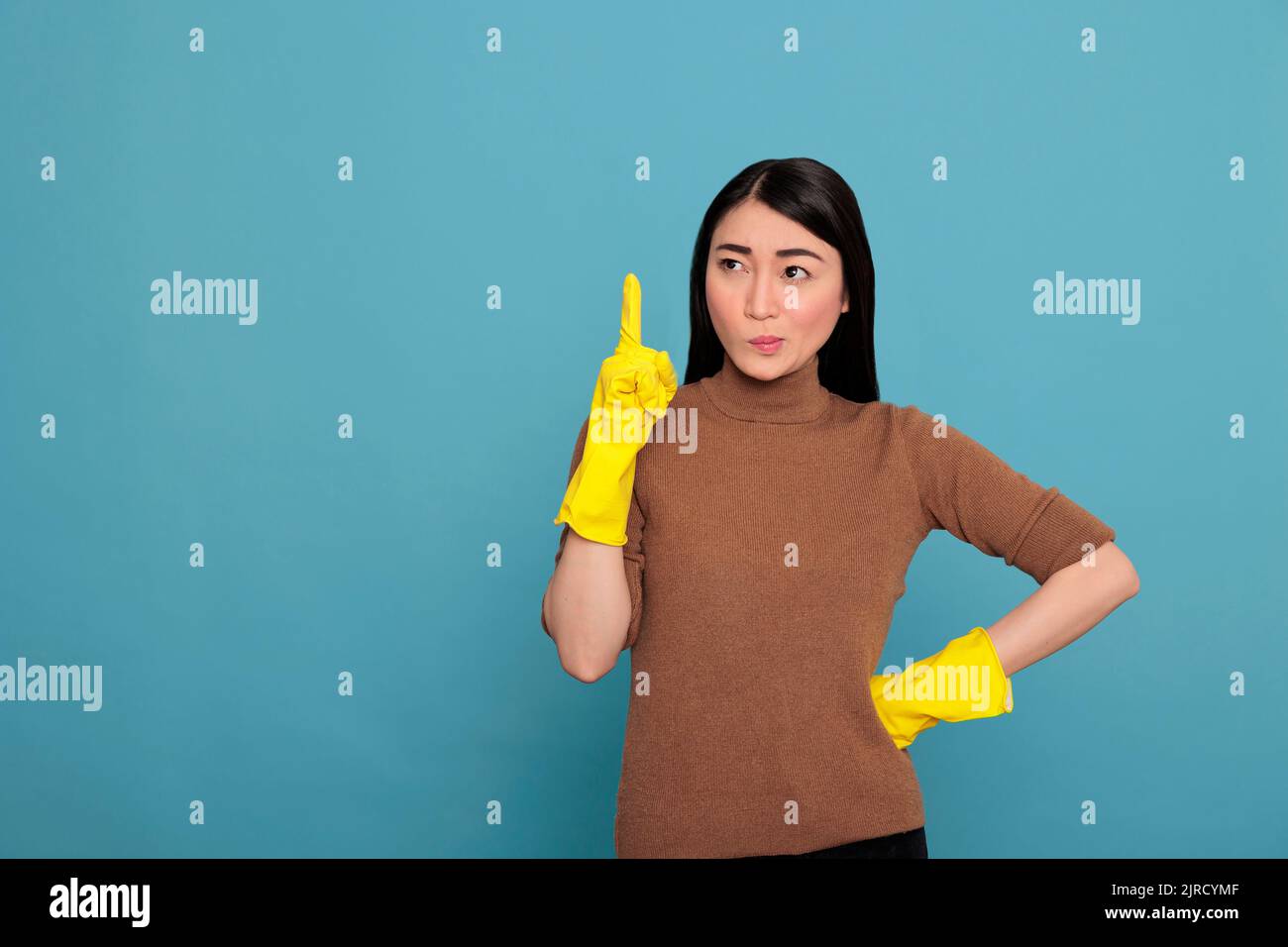 Young asian woman with contempt mood from chores wearing yellow gloves for hand safety and pointing finger upward look toward side isolated on a blue background, Cleaning home concept, Stock Photo