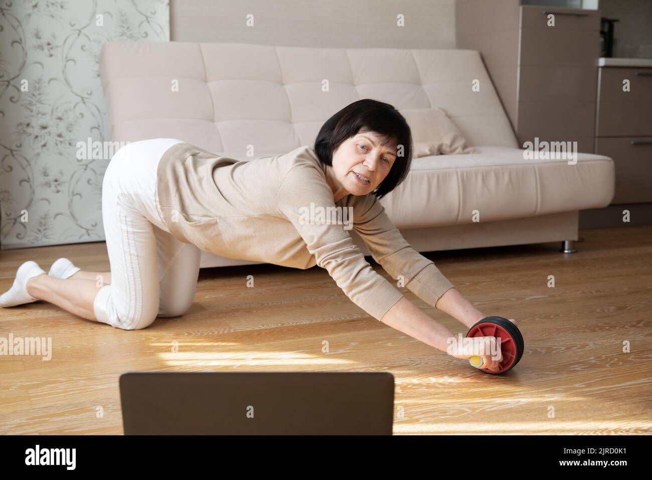 An active elderly woman does stretching exercises at home on the floor in front of a laptop. A mature lady takes care of her healthy body and spirit Stock Photo