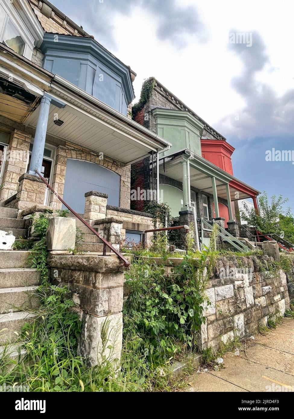 A few pairs of  charming 19th century vacant townhouses with large bay windows are boarded up and overgrown with weeds on a stormy summer day. Stock Photo
