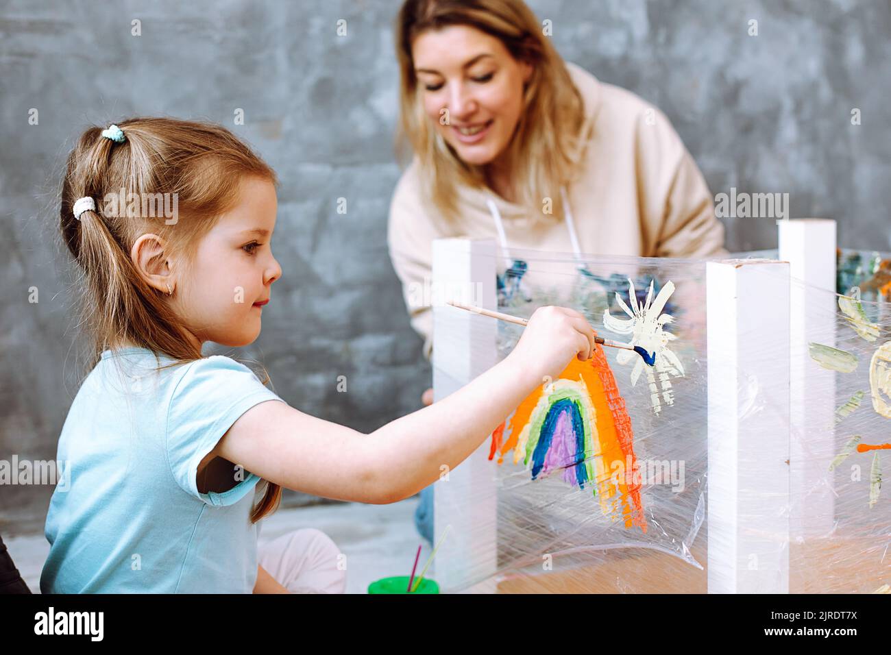Portrait of stunning little girl sit on art lesson drawing white sun near rainbow with brush on stretch film in room. Stock Photo