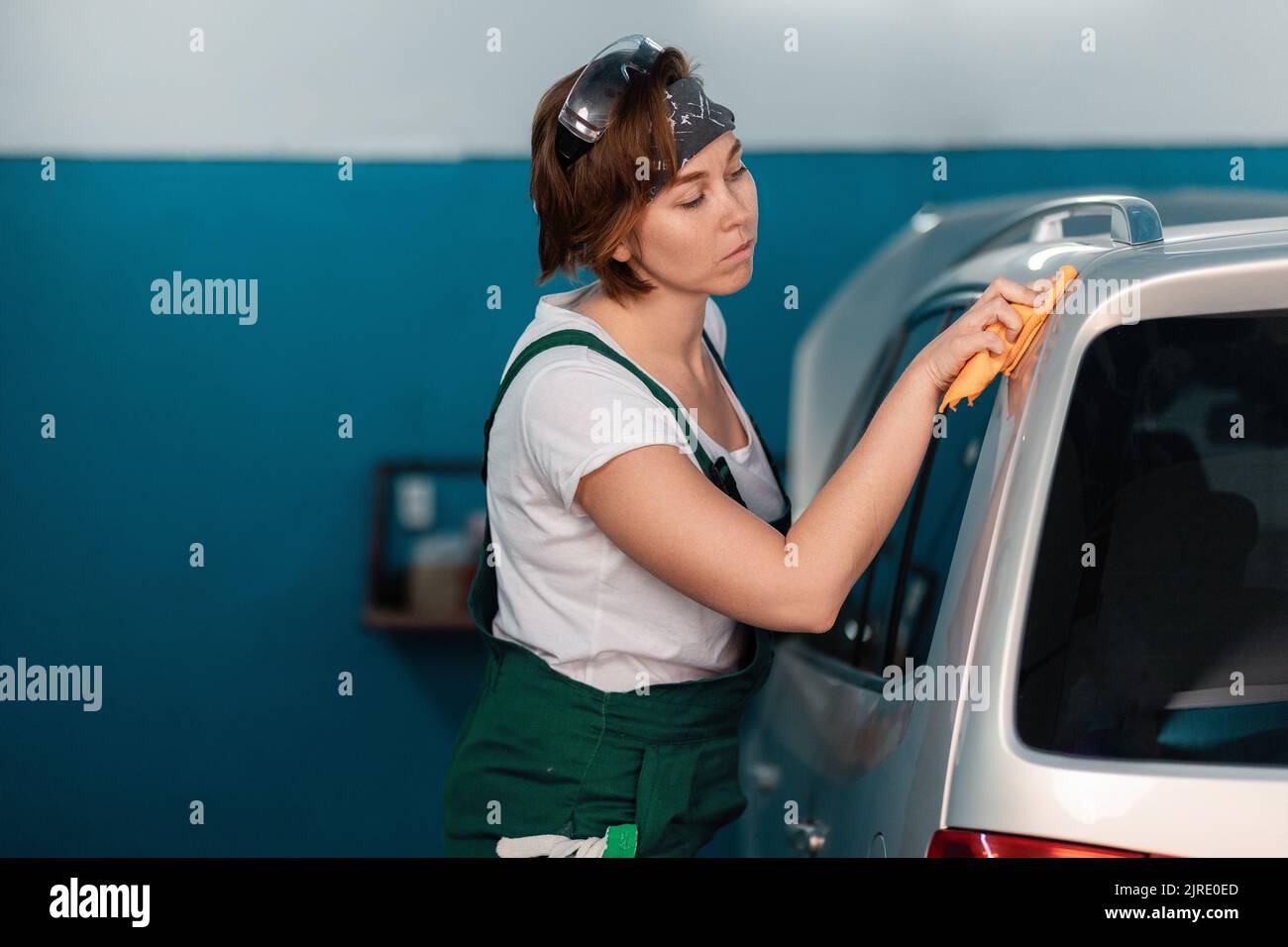 Caucasian young woman in a green coveralls wiping a car with a rag ...