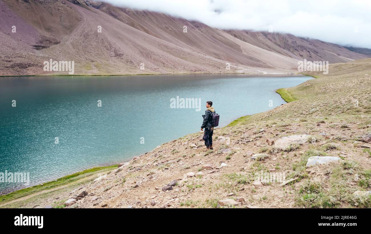 male tourist standing at Chandra Taal Lake overlooking blue glacier water in Spiti Valley India on cloudy day Stock Photo