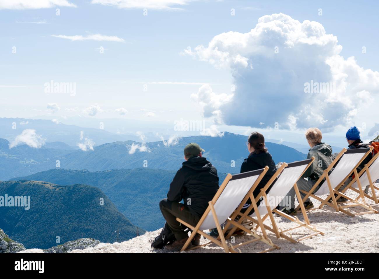 Group of people sitting in the deck chairs on a mountain top watching clouds above spectacular mountain view Stock Photo