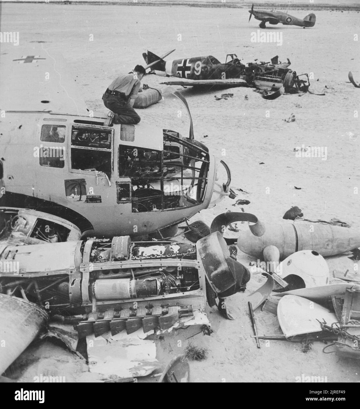 A vintage photo circa 1942 showing a British ground crewman inspecting a wrecked German Heinkel 111 bomber. A wrecked Messerschmitt BF109 aircraft is in the background along with a RAF Hurricane fighter near El Dabaa Egypt North Africa in World War Two Stock Photo