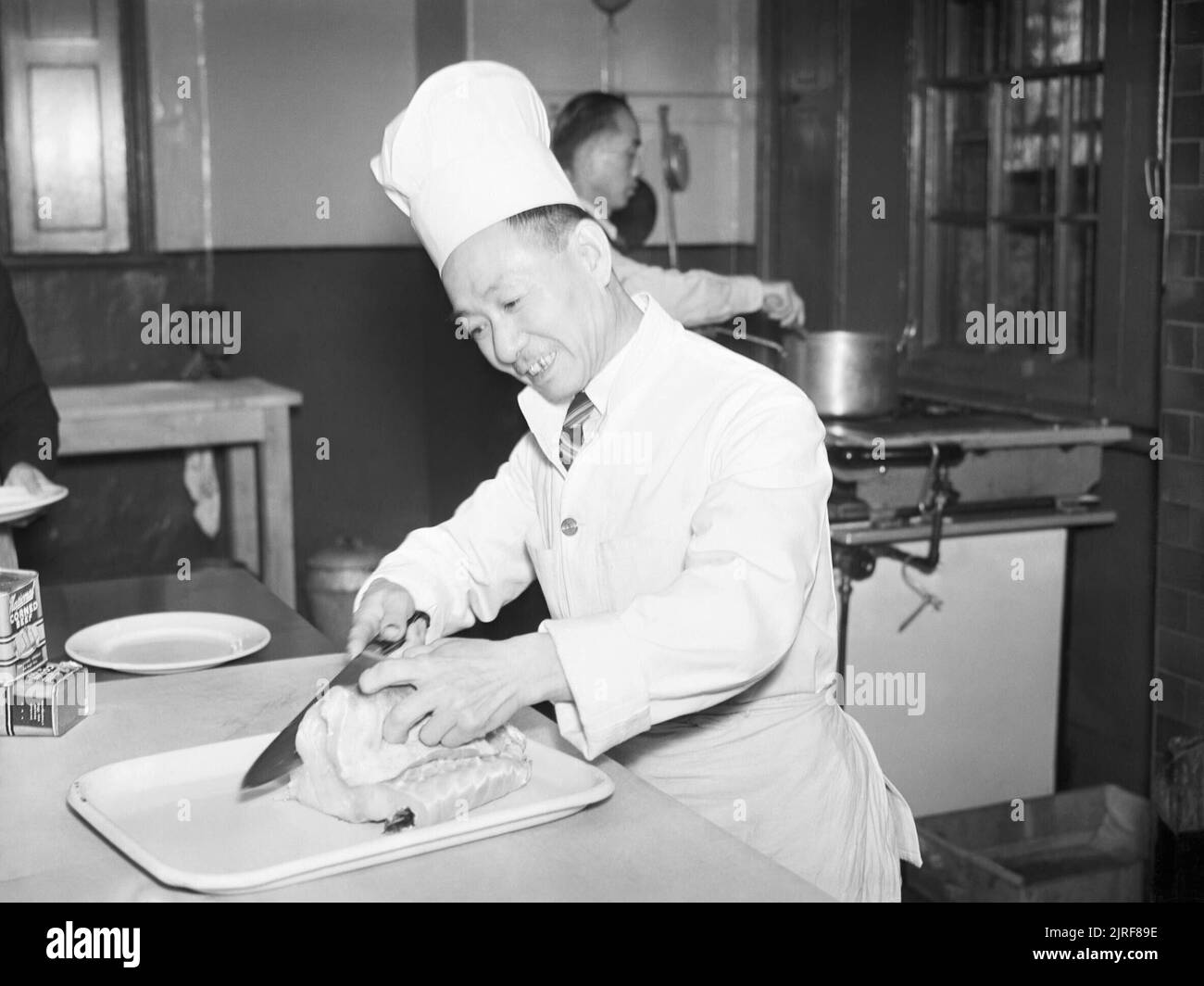 A Chinese merchant sailor, who served as chief cook, carves meat in the kitchen of the Chinese sailors' convalescent home in Liverpool, 1943. Ching Foo Po smiles as he carves meat in the kitchen of the Chinese convalescent home in Liverpool. According to the original caption, Ching Foo Po was a chief cook who came to the home after having suffered bad burns during a boiler explosion, but now cooks for the home as part of his convalescence. Stock Photo