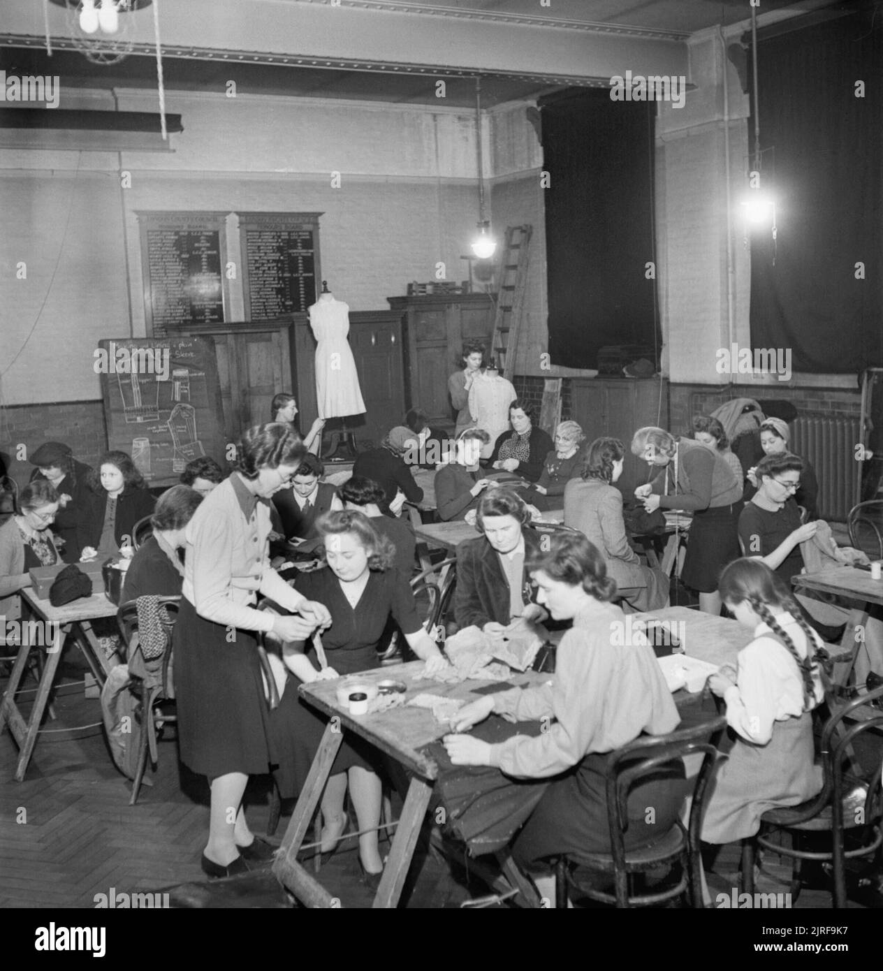 Mend and Make Do- Dressmaking Classes in London, 1943 Women sit at tables and work with needle and thread during a dressmaking class in a school in Brixton. Standing in the foreground, to help one of the women is Miss Clifford, a London County Council instructress. The LCC has been running such classes for many years, but their popularity has grown since the outbreak of the war, and the introduction of clothes rationing and Make Do and Mend. According to the original caption, Miss Clifford's jacket has been made out of a pair of white flannel trousers. One of the daughters of the women can als Stock Photo