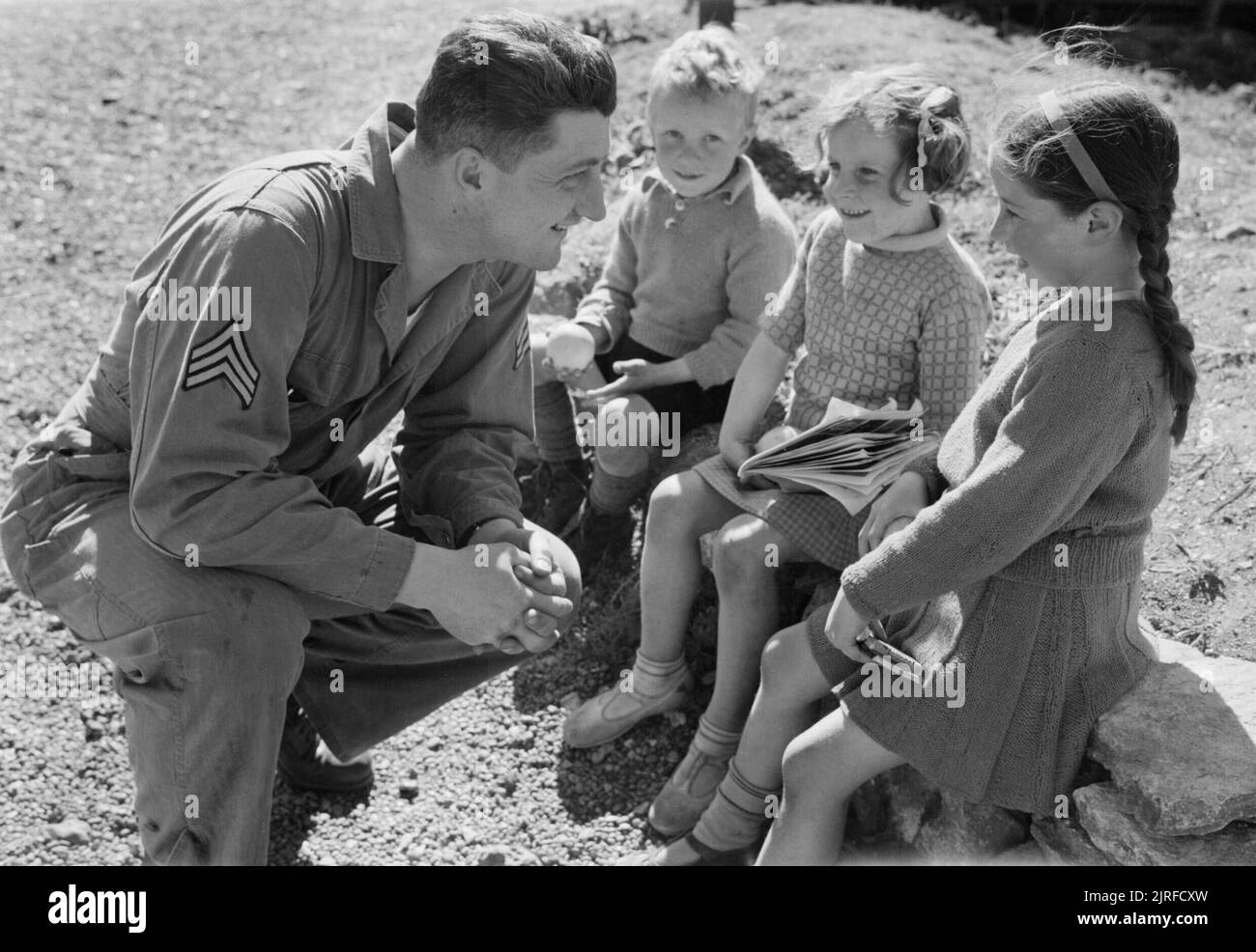 US Troops in An English Village- Everyday Life With the Americans in Burton Bradstock, Dorset, England, UK, 1944 Sergeant Foster Whitsitt (of Route 4, Box 915, Vancouver, Washington) smiles at three local children to whom he has just given oranges, in the sunshine in the village of Burton Bradstock, Dorset. The children include Chris Kerley and Betty 'Freckles' Mackay (right). Betty was given the nickname 'Freckles' by the Americans, as was a 'camp favourite'. Stock Photo