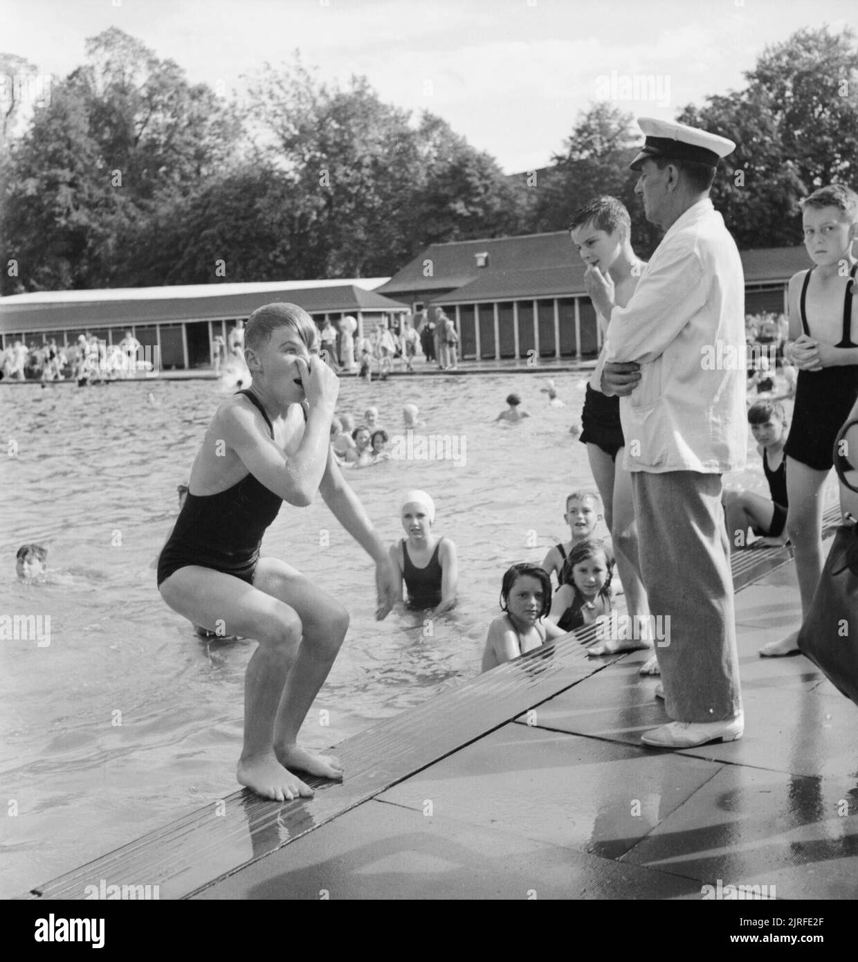 Bathing Pool- Entertainment and Relaxation in the Open Air, Guildford, Surrey, England, 1943 A young boy holds his nose tightly to prevent any water getting up his nostrils, as he prepares to jump backwards into the water at Guildford Lido. On the poolside, the guard or warden can be seen, overseeing the proceedings. A group of the boy's friends can be seen in the water, looking at the camera, enjoying a swim on a sunny afternoon. Stock Photo