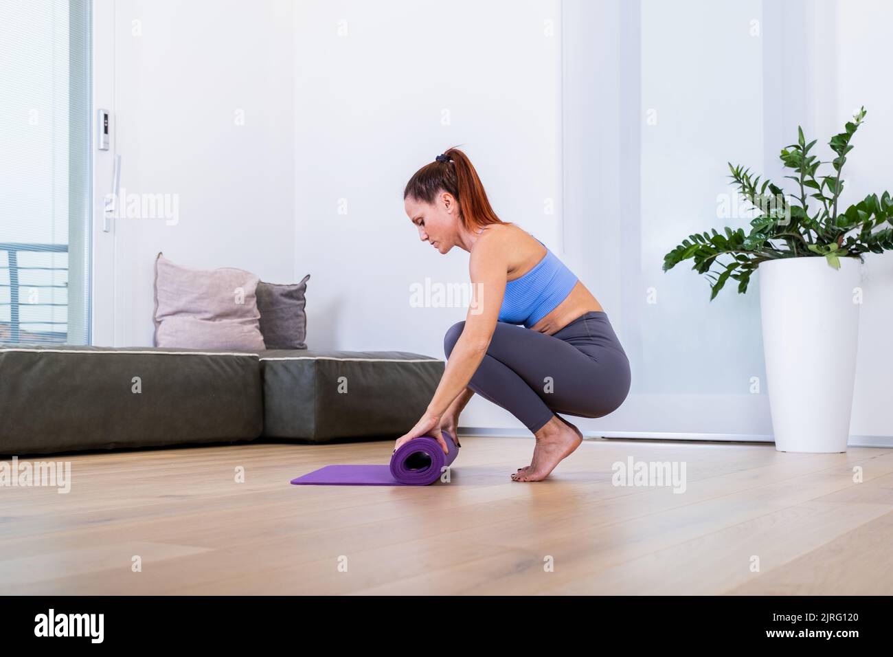Young yoga woman rolling her violet mat after a yoga class on wooden floor near a window in her linving room Stock Photo