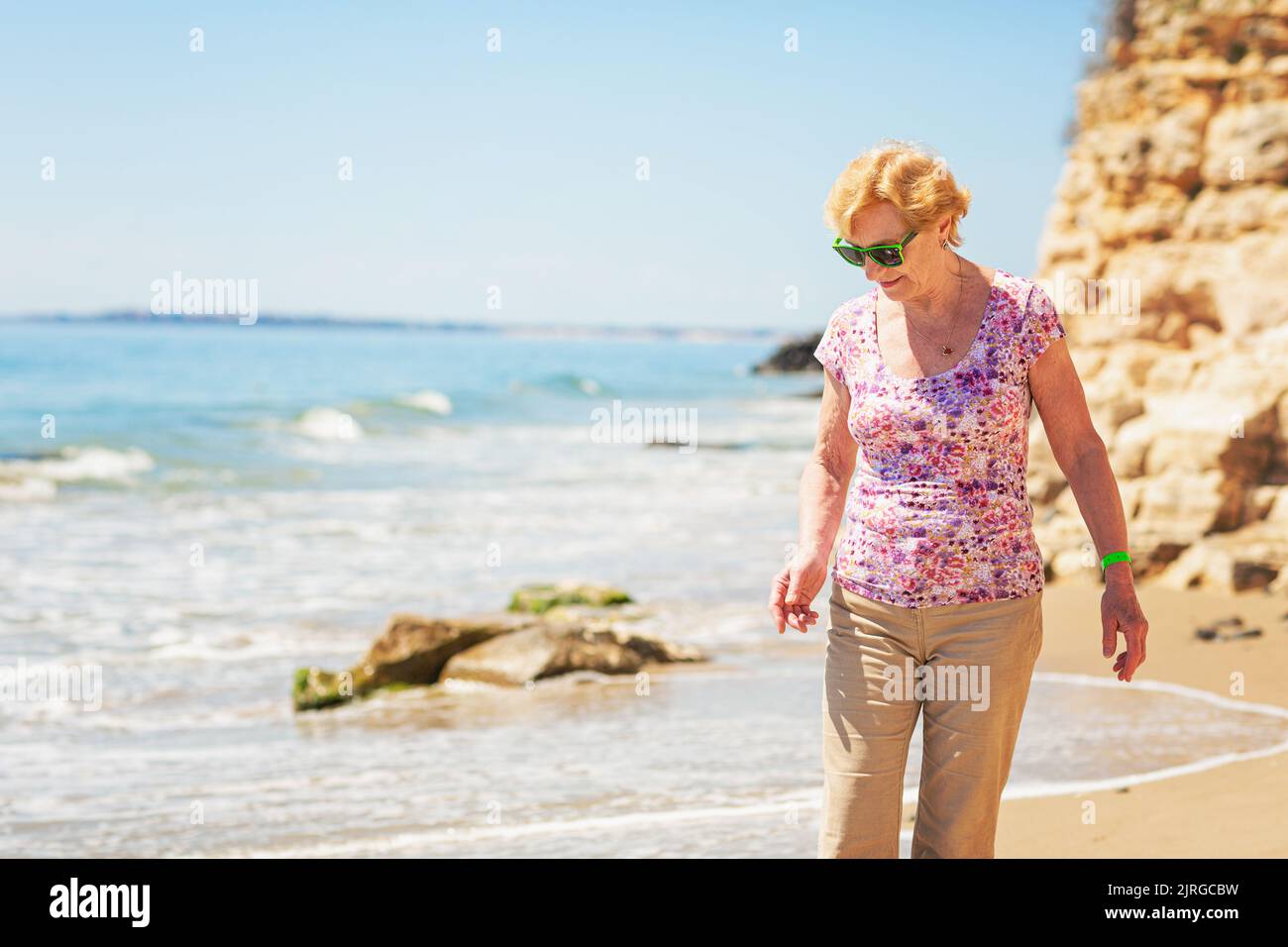 Senior woman in sunglasses stands on the beach and looks at the sea ...
