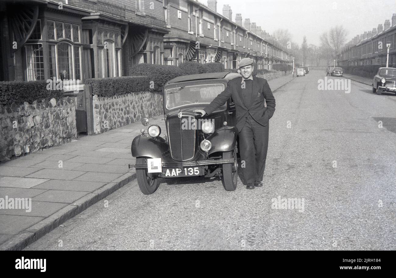1950s, historical, in a wide street of terraced houses, with decorative windows, England, UK, a man in a flat cap standing beside his small car, a pre-war Morris Eight. First released in1935, the Morris Eight became a popular small car, was in production until 1948 and saw Morris Motors become Britain's largest motor manufacturer. Stock Photo