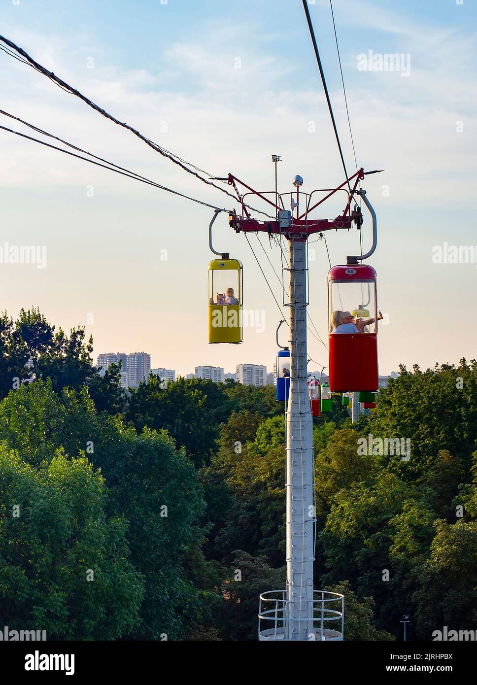 Colorful cableway over park with green trees at summer sunset, Kharkiv cityscape in background, Ukraine Stock Photo