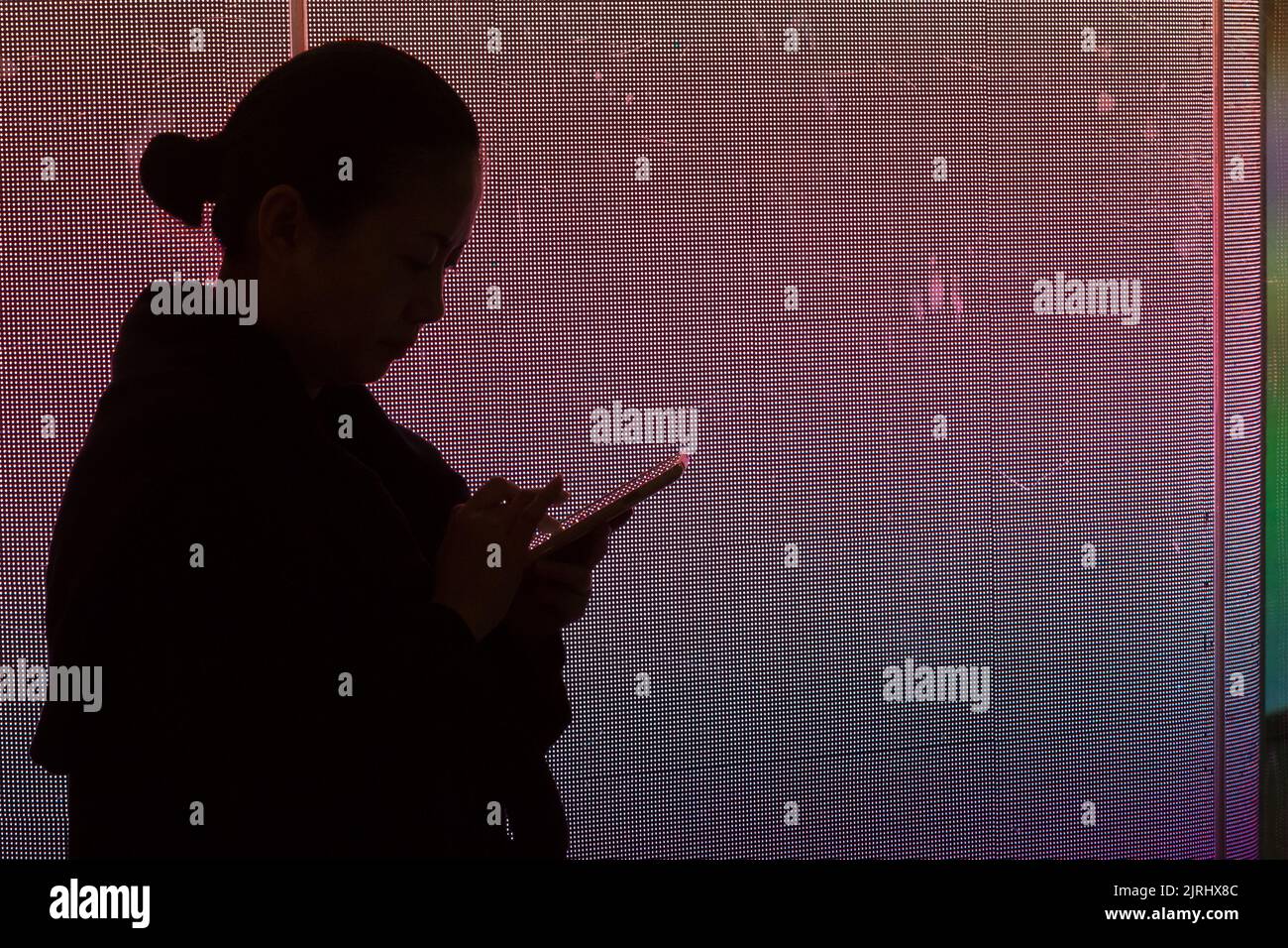 A woman, in silhouette, uses a smart phone against an illuminated wall. Shibuya, Tokyo, Japan. Stock Photo