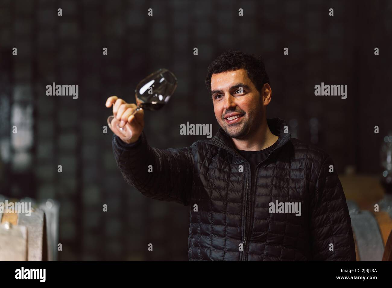 Man tasting red wine in a winery barrel cellar, holding a wine glass, swirling it, smelling, and sipping wine, delighted with wine taste and flavor. Stock Photo